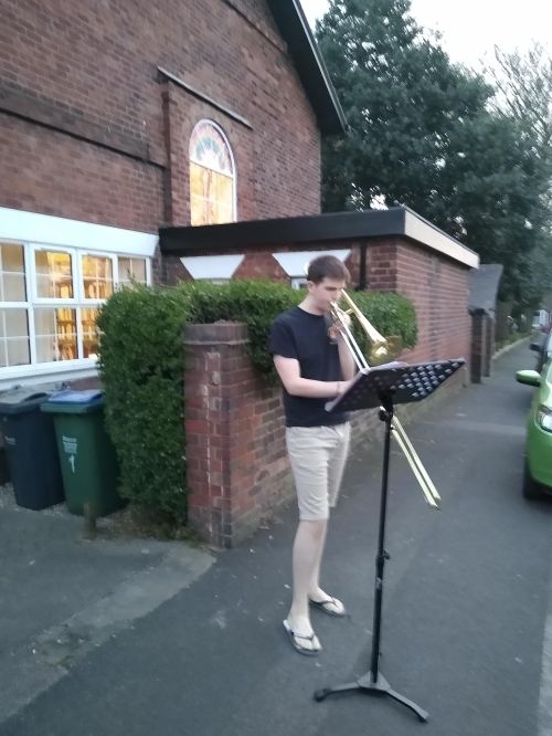 The Joker, wearing black t-shirt and beige shorts, flipflops, plays his trombone outside the Vicarage, using a black music stand.